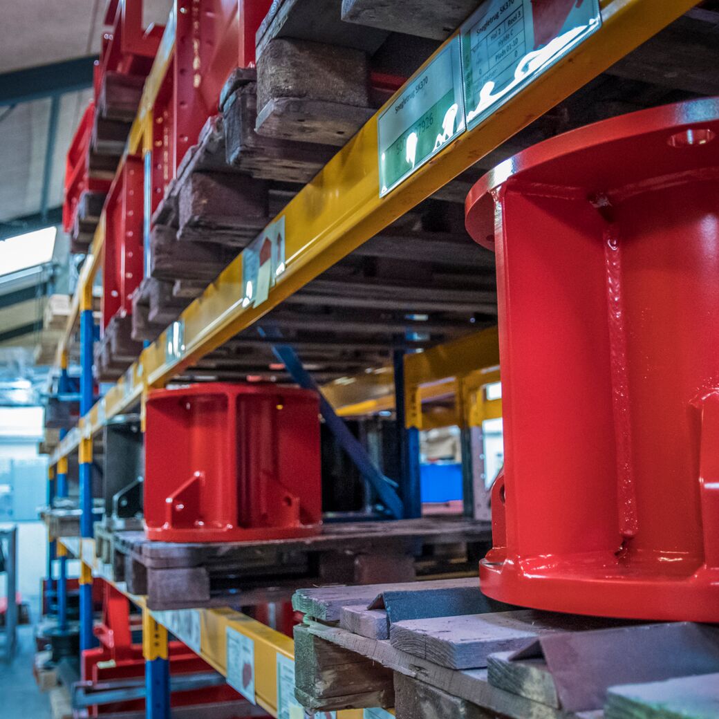 Metaldele opbevaret på paller i et industrielt reollager, med fokus på røde komponenter